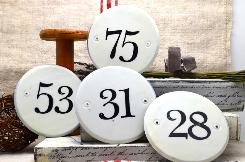 May include: Five white ceramic round and oval signs with black numbers. The numbers are 75, 53, 31, and 28.