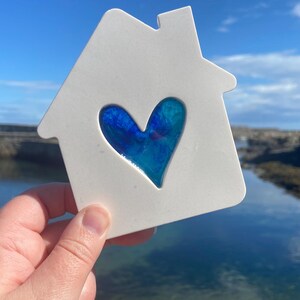 May include: White ceramic house shaped ornament with a blue and teal heart cutout. The heart is filled with a blue and teal glass piece.