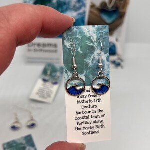 May include: Silver wave earrings with blue enamel, presented on a card with text about Portsoy, Scotland. The earrings feature a circular design with a wave motif. The card has a watercolour-style background.