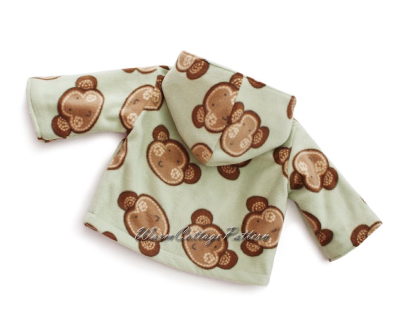 Op de afbeelding: Een lichtgroene fleece jas met capuchon voor een baby of peuter. De jas is bedekt met een herhalend patroon van bruine apengezichten. De tekst "Warm Cottage Pattern" staat op het onderste deel van de jas.