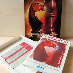 May include: A vintage box set for the Sargon III chess game for computers. The box is open and shows the game's manual and two floppy disks. The box features a red and brown chess knight and pawn on a red background.