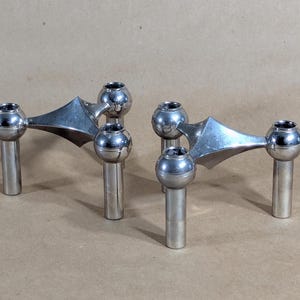 May include: Two silver-toned candle holders. Each holder has a central triangular design with three spherical candle cups and cylindrical bases. The candle holders are made of metal and have a polished finish. The design is modern and minimalist.