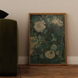 May include: A framed painting of white roses with green leaves on a light brown wood frame. The painting is hanging on a white wall above a green sofa with a black cushion.