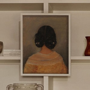 Op de afbeelding: Een wit rek met verschillende items erop, waaronder een ingelijst schilderij van een vrouw met donker haar, een witte keramische kan, een stapel boeken, een klein houten doosje en een bruine keramische vaas.