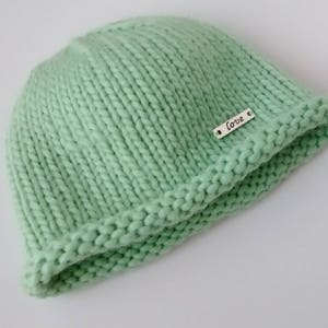 May include: A knitted beanie hat in a pale green colour with a turned-up brim. The hat features a small silver tag with the word "Love". The hat is made from a chunky knit.