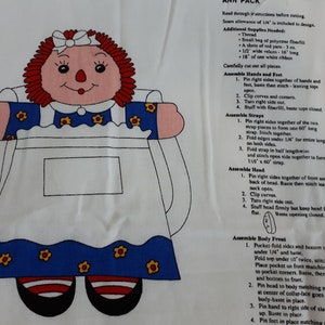 May include: A sewing pattern for a rag doll with instructions on how to assemble the doll. The doll is wearing a blue and white dress with red and white shoes. The pattern includes instructions for assembling the hands, feet, straps, head, and body.