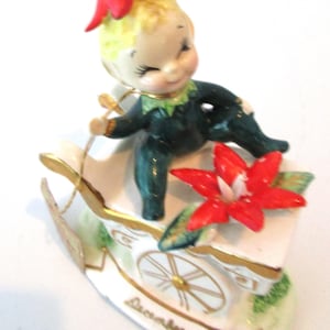 May include: A vintage ceramic figurine of a smiling elf in a green outfit and a red flower hat, seated on a white sleigh with gold accents. A red flower is on the sleigh, and the word "December" is written on it.