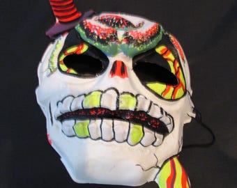May include: A white skull mask with black eye holes, yellow teeth, and red and yellow accents. A red and black striped dagger is attached to the top of the mask. The mask has a cracked appearance and is likely for Halloween or costume use.