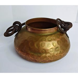 May include: A round, hammered copper pot featuring a wide opening and two dark, weathered iron ring handles attached to the sides. The metal surface displays a gradient of warm copper tones, ranging from bright metallic gold to deep reddish-brown.
