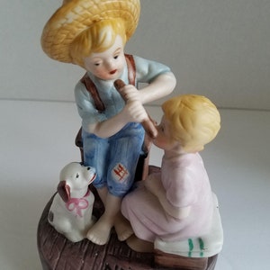 Puede incluir: Una figurita musical de porcelana que representa a dos niños, uno tocando la flauta para el otro. El niño lleva un sombrero de paja y un mono, y la niña lleva un vestido rosa. Un pequeño perro blanco con un lazo rosa está sentado en la base. La base es marrón y tiene las palabras "Norman Rockwell" inscritas en ella.