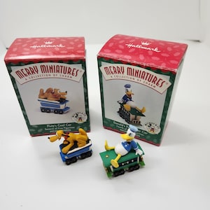 May include: Two Hallmark Merry Miniatures boxes with Pluto and Donald Duck figurines. Pluto is in a blue coal car, and Donald Duck is in a green coal car. The boxes are red with green trim and the words "Merry Miniatures" on them.