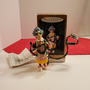 May include: A Christmas ornament featuring a woman wearing a colorful dress and sunglasses, holding a string of Christmas lights. The ornament is part of a Hallmark Keepsake Ornament collection.