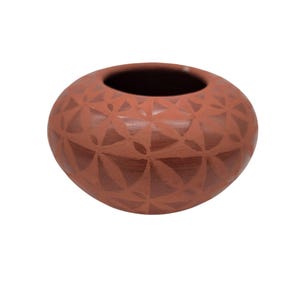 Op de afbeelding: Een terracotta-kleurige, ronde aardewerken kom met een brede opening. De buitenkant heeft een herhalend geometrisch patroon van driehoeken en gebogen lijnen. De kom is waarschijnlijk handgemaakt en geschikt voor decoratieve doeleinden.