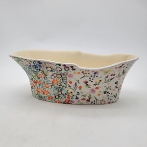 Puede incluir: Una jardinera ovalada de cer&aacute;mica con interior crema y un estampado floral en el exterior. El dise&ntilde;o floral presenta una variedad de flores coloridas, incluyendo naranjas, azules y moradas, sobre un fondo crema. La jardinera tiene una parte superior ancha y abierta.