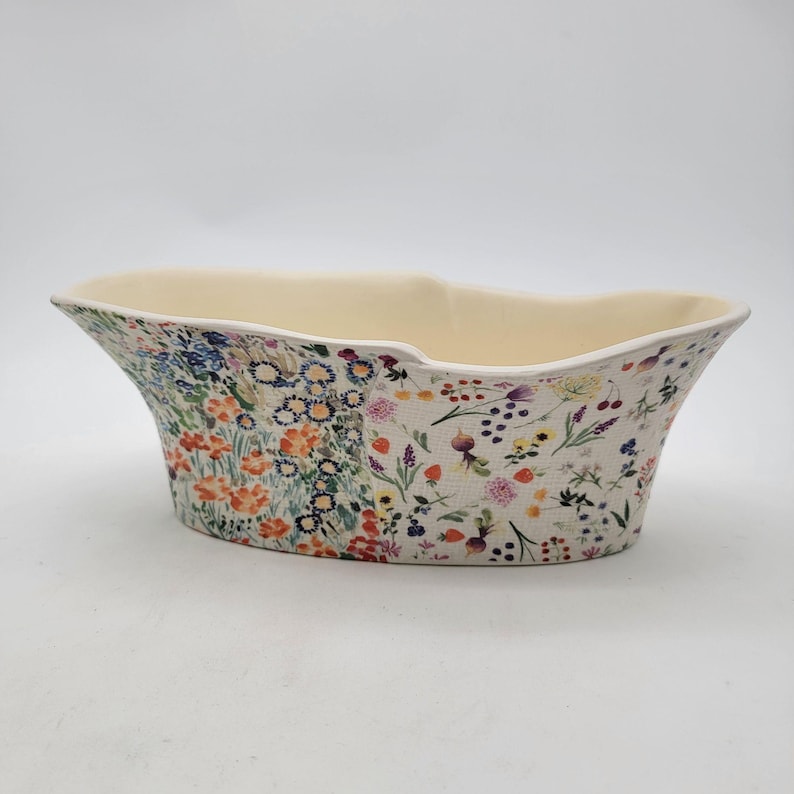 Puede incluir: Una jardinera ovalada de cer&aacute;mica con interior crema y un estampado floral en el exterior. El dise&ntilde;o floral presenta una variedad de flores coloridas, incluyendo naranjas, azules y moradas, sobre un fondo crema. La jardinera tiene una parte superior ancha y abierta.