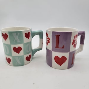 May include: Two ceramic mugs. One mug has a teal and white checkerboard pattern with red hearts and the letters X and O. The other mug has a purple and white checkerboard pattern with a red heart and the letters L and E.