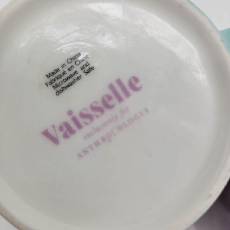 May include: White ceramic mug with the word "Vaisselle" in pink lettering. The mug has a sticker that says "Made in China, Microwave and dishwasher safe."