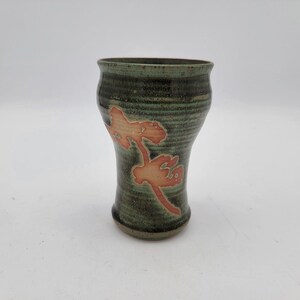 May include: A cylindrical ceramic tumbler featuring a dark forest green glaze with horizontal ridged textures. The exterior surface is decorated with a prominent, abstract orange-red floral or botanical motif that contrasts against the dark base.