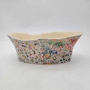 Puede incluir: Una jardinera ovalada de cer&aacute;mica con interior crema y un dise&ntilde;o floral en el exterior. El patr&oacute;n presenta una variedad de flores y follaje coloridos sobre un fondo claro. La jardinera tiene un borde ondulado.