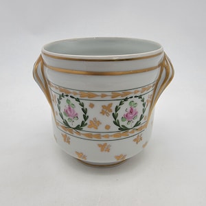 May include: A white porcelain vessel with two side handles, featuring a hand-painted design of pink roses within green laurel wreaths, accented by gold leaf trim and small gold floral motifs around the body and along the upper rim.