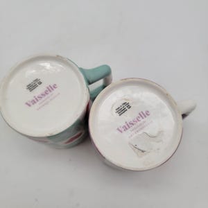 May include: Two ceramic mugs with the brand name "Vaisselle" printed on the bottom. One mug has a mint green handle and a pattern of red and white shapes. The other mug has a white handle and a purple rim.