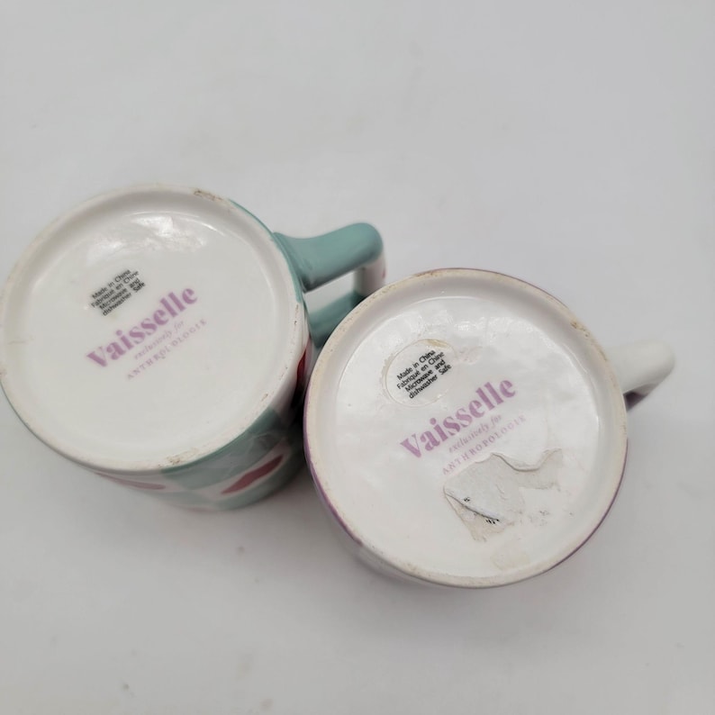 May include: Two ceramic mugs with the brand name "Vaisselle" printed on the bottom. One mug has a mint green handle and a pattern of red and white shapes. The other mug has a white handle and a purple rim.