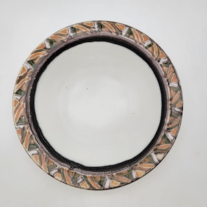 Op de afbeelding: Een ronde keramische kom met een witte binnenkant en een decoratieve rand. De rand heeft een herhalend patroon van oranje, groene en witte geometrische vormen tegen een zwarte rand. De kom is gecentreerd op een witte achtergrond.