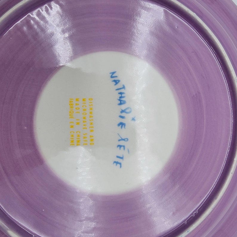 Puede incluir: Un plato de cer&aacute;mica con un centro blanco y un borde lavanda. El plato tiene el texto "Nathalie Rete" en escritura azul. El plato est&aacute; marcado como "Dishwasher and Microwave Safe Made in China Fabriqu&eacute; en Chine".