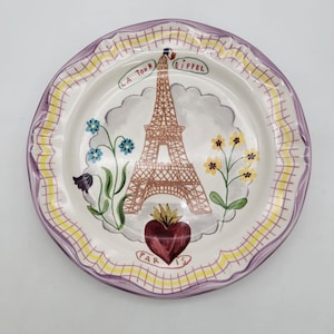 Puede incluir: Un plato de cer&aacute;mica decorativo con un dise&ntilde;o pintado a mano de la Torre Eiffel. El plato tiene un centro blanco con una torre marr&oacute;n, flores azules y amarillas y un coraz&oacute;n rojo. El borde es morado y amarillo.