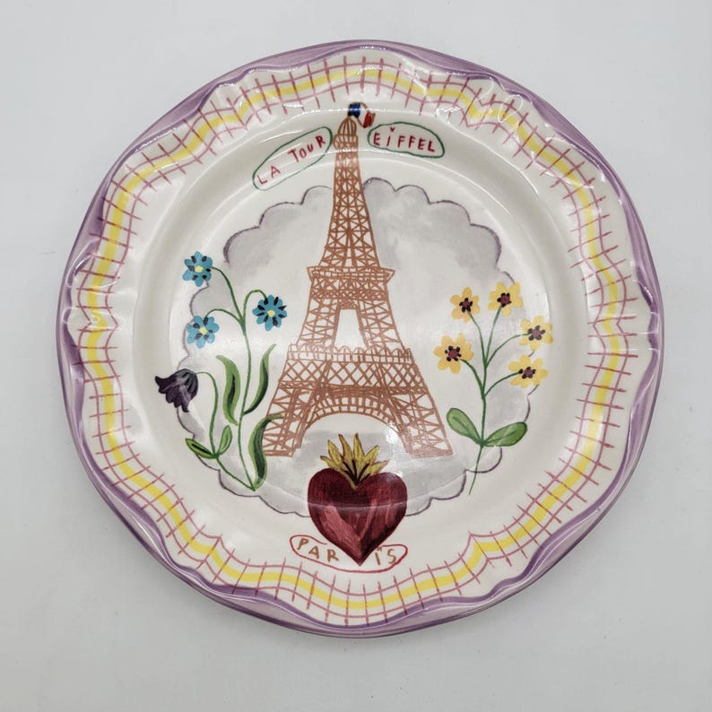 Puede incluir: Un plato de cer&aacute;mica decorativo con un dise&ntilde;o pintado a mano de la Torre Eiffel. El plato tiene un centro blanco con una torre marr&oacute;n, flores azules y amarillas y un coraz&oacute;n rojo. El borde es morado y amarillo.
