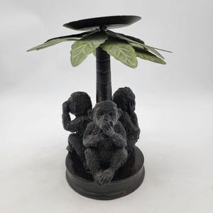 Può includere: Un portacandele marrone scuro con tre statuette di scimmie nella posa 'non vedere il male, non sentire il male, non parlare il male'. Le scimmie sono posizionate attorno a un tronco di palma centrale, sormontato da una piattaforma per candele nera e foglie di palma verdi.