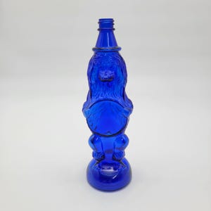 May include: A cobalt blue glass bottle shaped like a standing poodle with a textured coat, a smooth circular area on the chest, and a narrow neck with a threaded top. The figure stands on a circular base, featuring moulded details of the dog's fur.