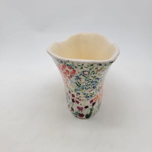 Puede incluir: Un jarr&oacute;n de cer&aacute;mica con un estampado floral. El jarr&oacute;n tiene un interior color crema y un borde acampanado y ondulado. El exterior est&aacute; decorado con un colorido patr&oacute;n de flores rojas, azules y moradas y hojas verdes sobre un fondo blanco.