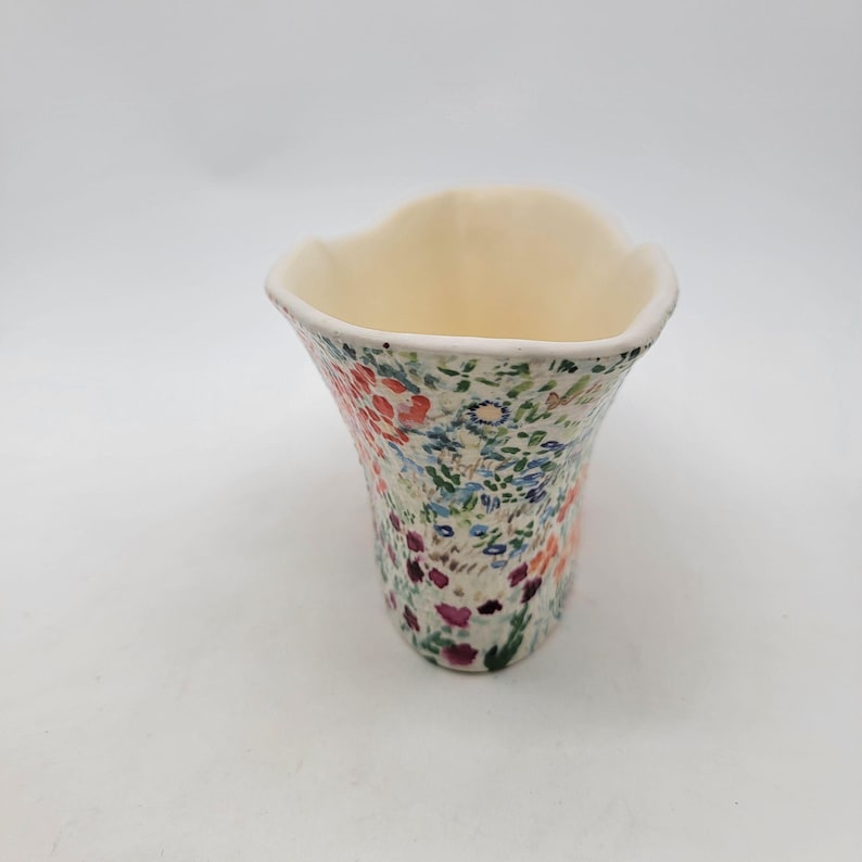 Puede incluir: Un jarr&oacute;n de cer&aacute;mica con un estampado floral. El jarr&oacute;n tiene un interior color crema y un borde acampanado y ondulado. El exterior est&aacute; decorado con un colorido patr&oacute;n de flores rojas, azules y moradas y hojas verdes sobre un fondo blanco.