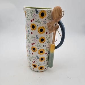 May include: A white ceramic pitcher with a sunflower pattern, a blue handle, and a green rim. A wooden spoon with a yellow and green handle is tied to the pitcher with twine. The pitcher is approximately 9 inches tall.