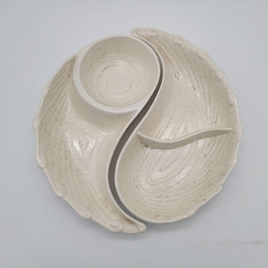 May include: Cream-colored ceramic serving dish with a central bowl and two curved compartments. The dish has a textured surface with speckled details and a leaf-like design. The bowl is round with a spiral pattern.