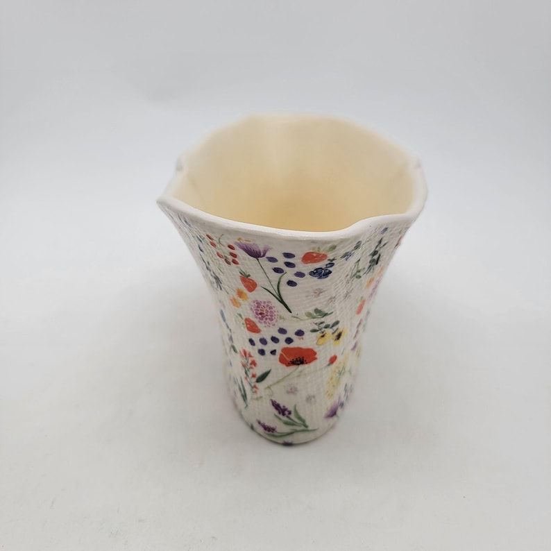 Puede incluir: Un jarr&oacute;n de cer&aacute;mica color crema con un estampado floral. El jarr&oacute;n tiene forma acampanada y un borde con volantes. Las flores son en tonos rojos, morados, naranjas y amarillos.