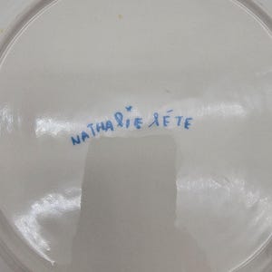 Puede incluir: Un plato de cer&aacute;mica blanco con la firma manuscrita azul "Nathalie Lete". El plato tiene un acabado brillante y un borde ligeramente curvado. La firma es de estilo cursivo.