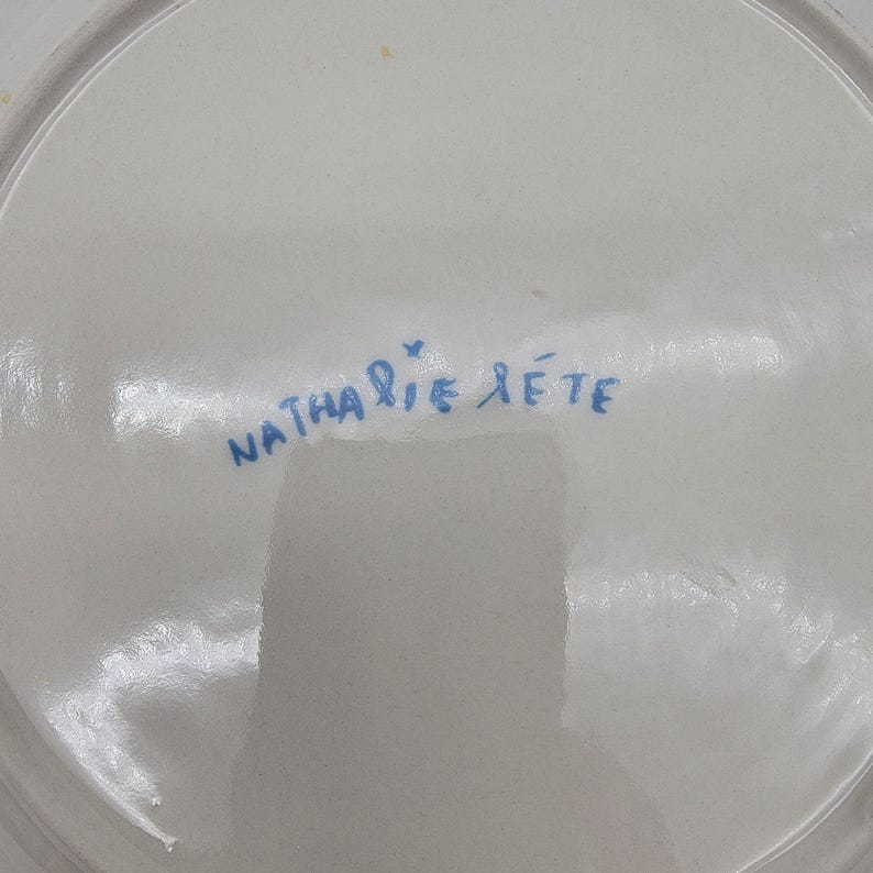 Puede incluir: Un plato de cer&aacute;mica blanco con la firma manuscrita azul "Nathalie Lete". El plato tiene un acabado brillante y un borde ligeramente curvado. La firma es de estilo cursivo.