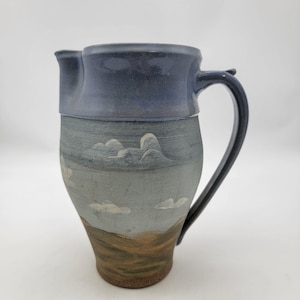 Może przedstawiać: Ręcznie robiony ceramiczny dzbanek z niebieskim wierzchem i uchwytem oraz szarym korpusem. Korpus zdobi krajobraz z białymi chmurami i brązowymi wzgórzami. Dzbanek posiada dzióbek.