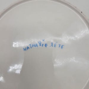 以下が含まれることがあります： 青色の手書き文字「Nathalie L&eacute;t&eacute;」が入った白いセラミックプレート。 プレートは光沢仕上げで、縁がわずかに湾曲しています。 テキストは中央に配置されており、署名のように見えます。