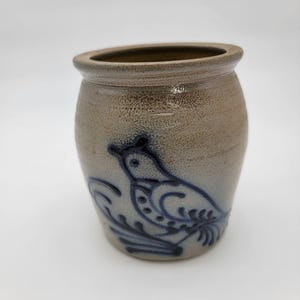 May include: A tan stoneware crock featuring a blue cobalt slip-trailed bird design on the front. The vessel has a thick, rounded rim and a stamped oval maker's mark near the top edge that includes the text York, Maine, visible on the surface.