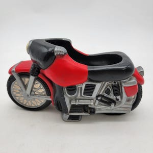 Op de afbeelding: Een rode, zwarte en zilverkleurige motorfietsvormige plantenbak. De plantenbak heeft een gedetailleerd ontwerp, inclusief wielen, een zitting en motoronderdelen. De motorfiets heeft een zwarte zitting en een rode carrosserie. De wielen zijn zilver.