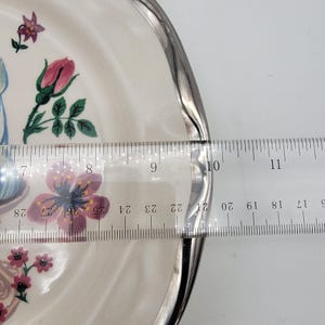 以下が含まれることがあります： ピンク、紫、緑の花柄で装飾された、銀色の縁が付いた白いセラミックプレート。 プレートの直径は約23cmです。