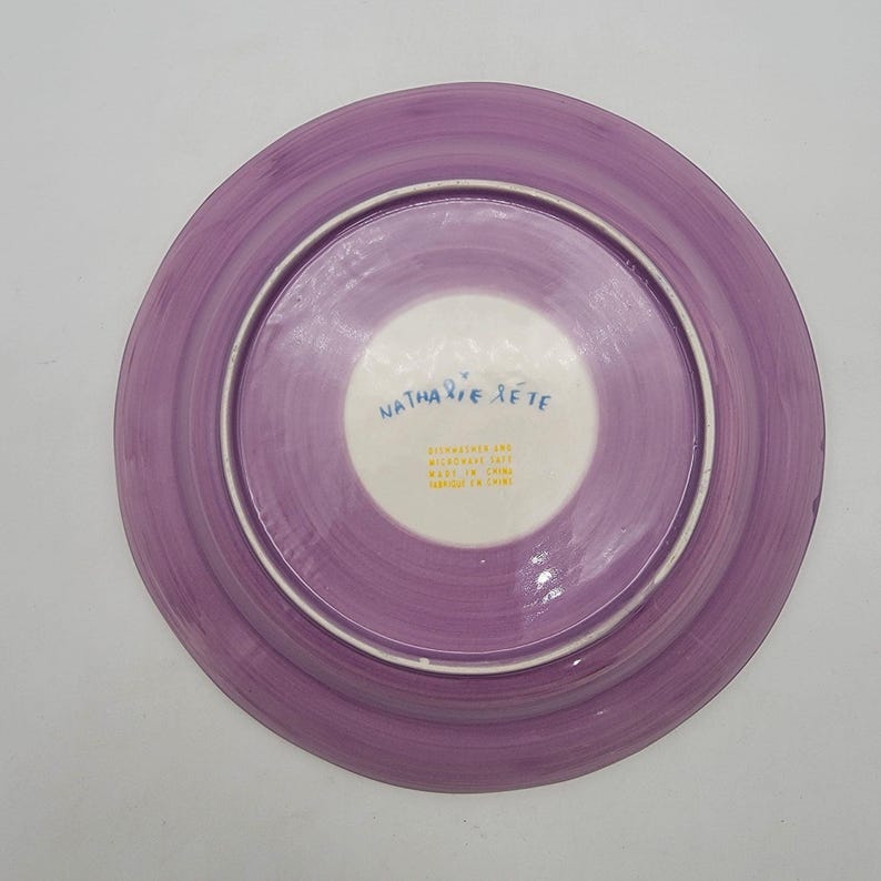 Puede incluir: Un plato de cer&aacute;mica con borde morado y centro blanco. El plato tiene un aspecto pintado a mano con pinceladas visibles. El texto "Nathalie Let&eacute;" est&aacute; escrito en azul en el centro blanco. El plato es apto para lavavajillas y microondas.