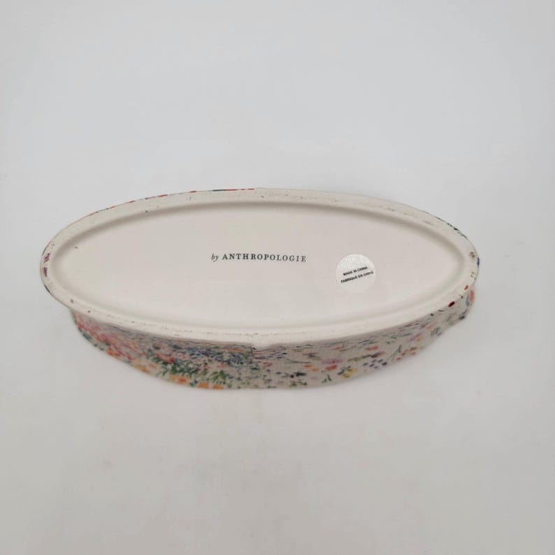 Puede incluir: Un plato de cer&aacute;mica ovalado con un estampado floral. El plato tiene un interior blanco y un exterior texturizado con un dise&ntilde;o floral colorido. Las palabras "by ANTHROPOLOGIE" est&aacute;n impresas en la parte inferior.
