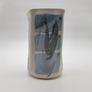 May include: A cylindrical ceramic pitcher with an off-white base glaze. The front features three stacked light blue rectangular patches, each overlaid with a dark gray, sweeping brushstroke design and a single small black dot.