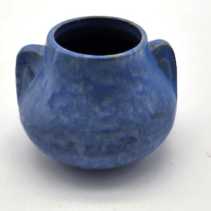 May include: A rounded ceramic vessel featuring a vibrant blue glaze with subtle tonal variations and a crackled texture. The piece includes two integrated side handles and a wide, circular opening at the top, showcasing a deep, dark interior.