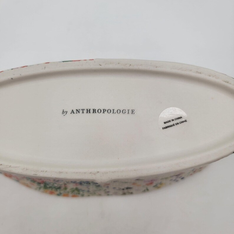 Puede incluir: Un plato ovalado de cer&aacute;mica blanca con un estampado floral en el borde. La base del plato tiene el texto "by ANTHROPOLOGIE" y una pegatina que dice "MADE IN CHINA / FABRIQU&Eacute; EN CHINE".
