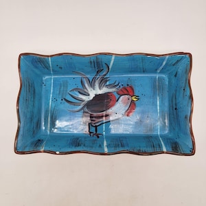 May include: Rectangular ceramic serving dish with a turquoise background and a hand-painted rooster design. The rooster is depicted with red, white, black, and yellow accents. The dish has a wavy brown rim and white vertical stripes.